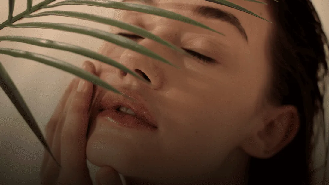 A woman resting her hand against her cheek with her eyes closed, a palm frond crossing the frame.